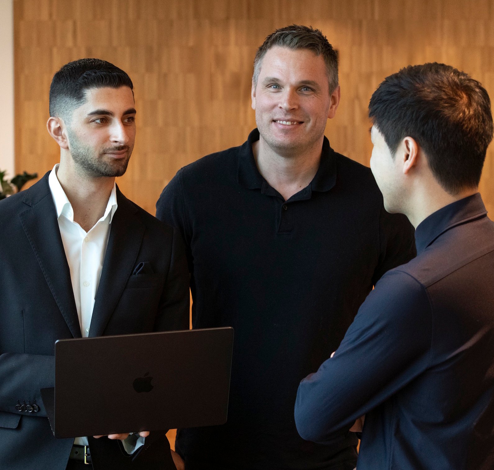 Tre män som står och pratar med varandra, varav en håller en dator i handen