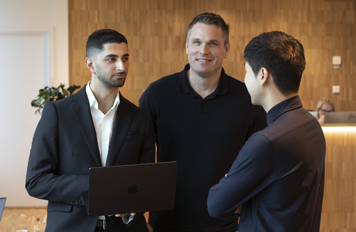 Tre män som står och pratar med varandra, varav en håller en dator i handen